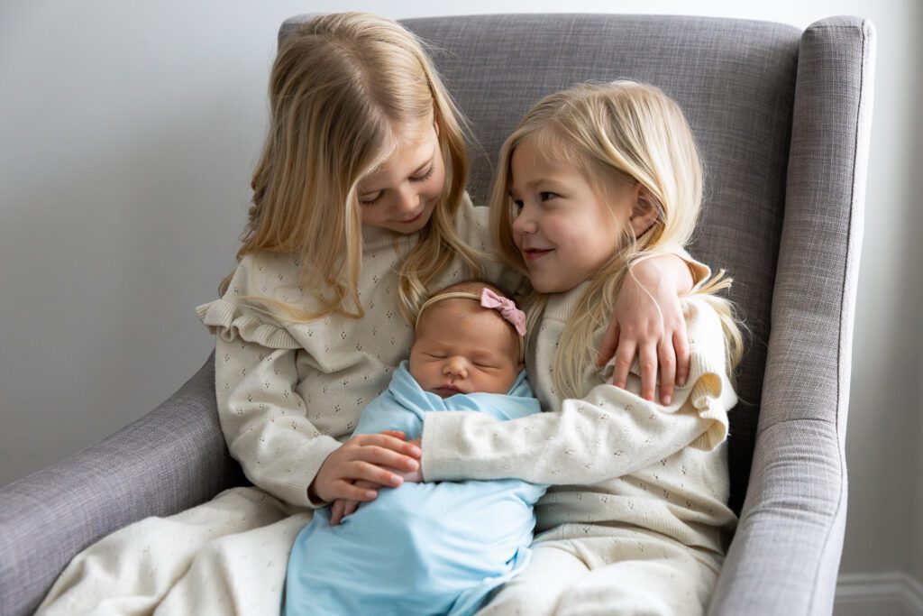 Two sisters hold newborn sister on chair in Roswell